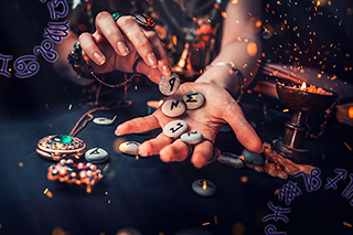 Psychic intuitive places a collection of rune stones in her hand with sparks of fire in the air against a dark background.